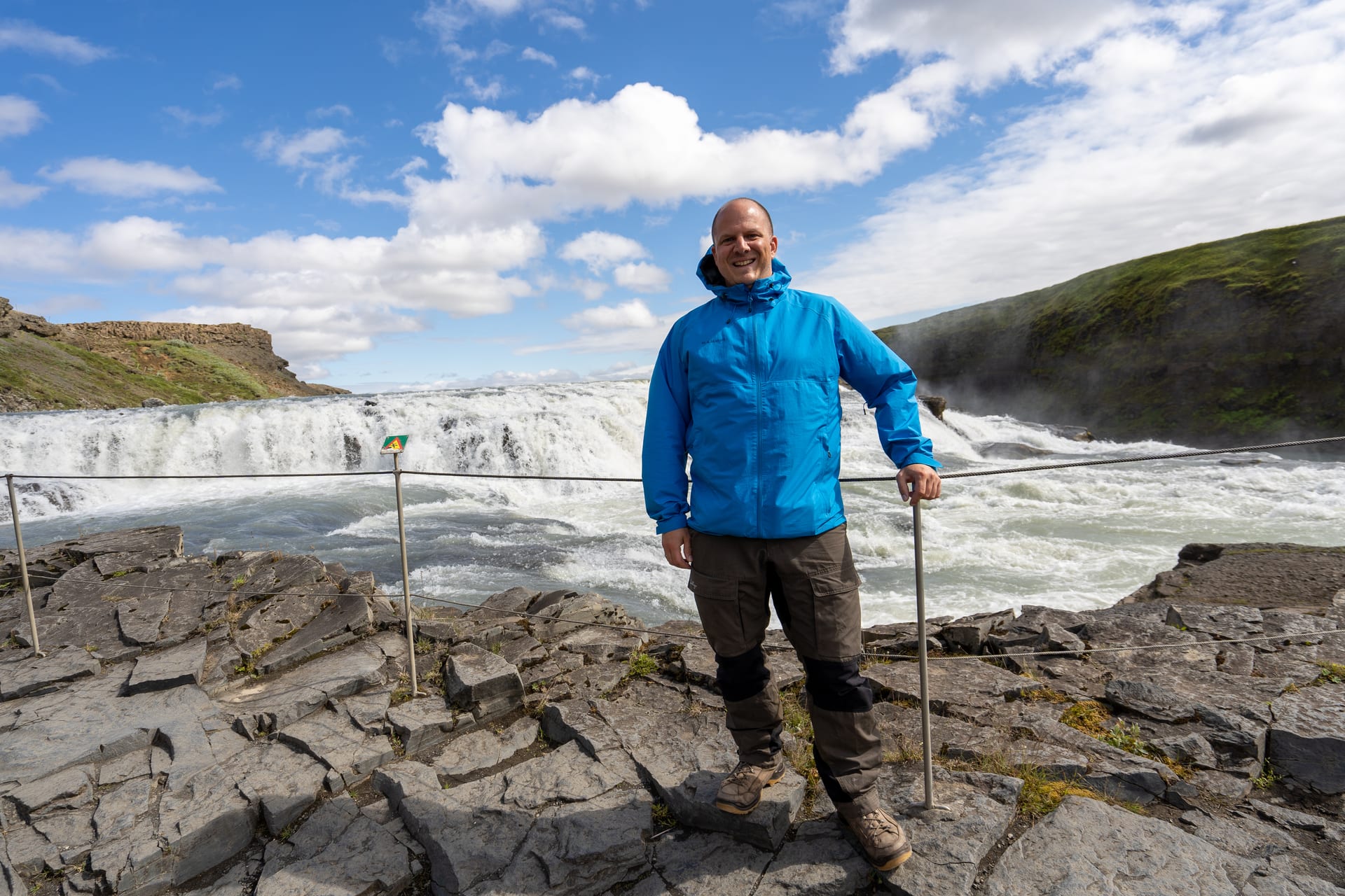 Mann in blauer Jacke vor dem zweistufigen Gullfoss-Wasserfall in Island, blauer Sommerhimmel mit Wolken.